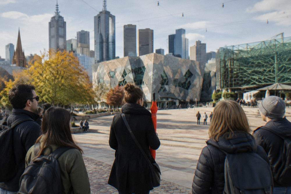 A guide holding an orange umbrella with a small tour group gathered on the steps of Federation Square in Melbourne