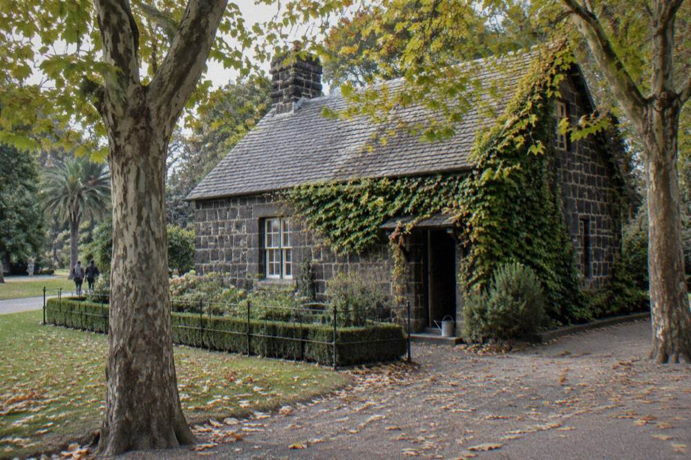 A classical stone cottage with a tidy garden surrounded by large green trees