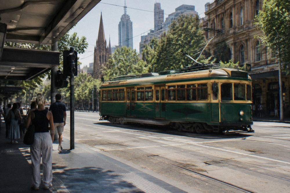 A green and gold Melbourne tram crossing a busy CBD intersection with pedestrians waiting to board