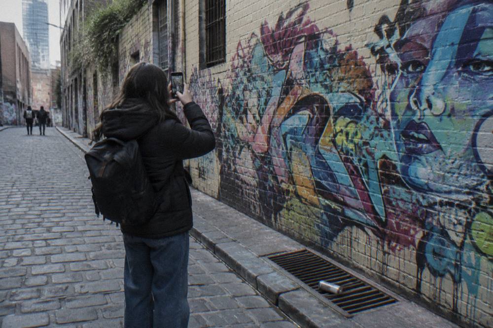A visitor photographing dense layered graffiti along a cobbled laneway wall